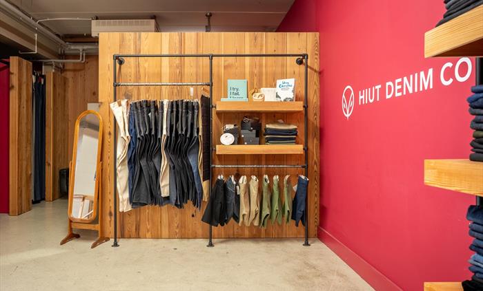 Hiut Denim Factory shop display with folded jeans, fabric swatches, and books on wooden shelves against a red wall. A full-length mirror and fitting r